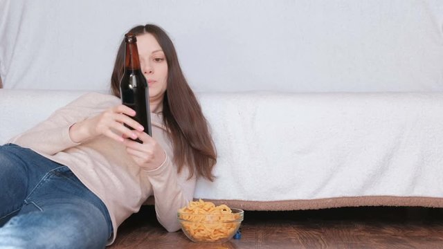Young beautiful woman drinking a beer and eating chips laying on the floor near the couch. Reads the label on the bottle and surprised.