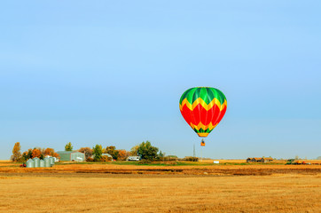 A multi-colored balloon is flying over a field in the sky near a farm