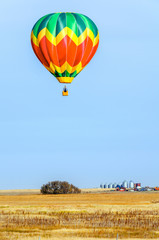 A multi-colored balloon is flying over a field in the sky near a farm