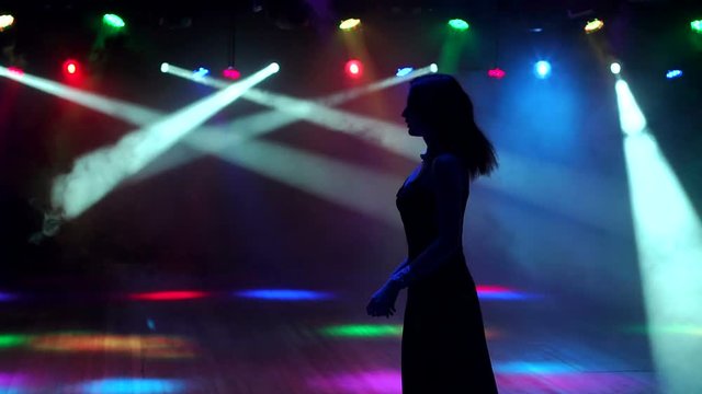 Cute beautiful brunette in evening dress dancing at a bachelorette party on the background of bright spotlights. Silhouette. Slow motion.