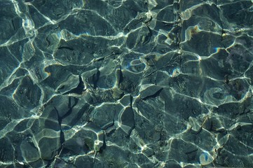 Horizontal photo of cristal clear water with rocks and sand at the bottom and sunlight reflections