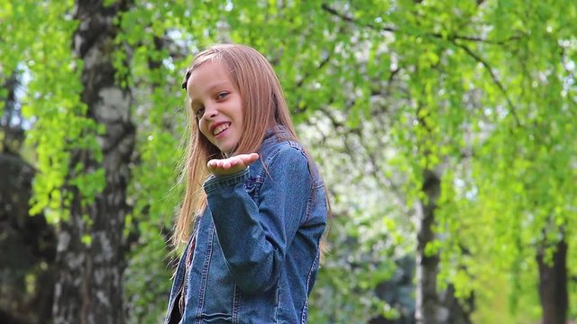 Portrait of pretty blonde teenage girl blowing kiss into camera. Teenager standing in green city park outdoors on warm spring day.