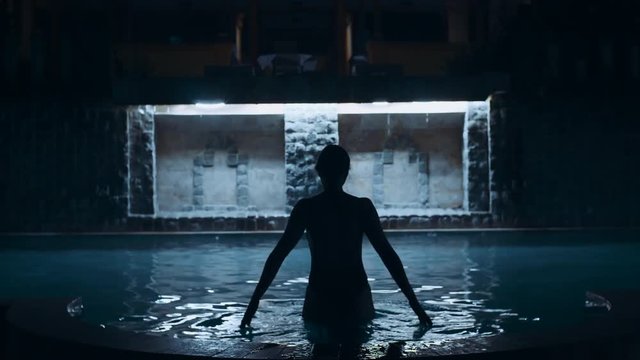 Night footage of beautiful young girl with amazing athletic sexy body in black bikini comes into the water and swims in the swimming pool with light, old vintage wall. Movie shooting of female model