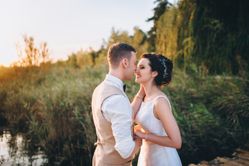 Lovely bride and groom embrace and smile at each other against the background of reeds and a lake. Newlyweds at sunset. Summer wedding.
