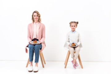Naklejka premium mother and daughter sitting on chairs and holding plates with pancakes on white