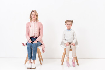 Naklejka premium mother and daughter sitting on chairs and looking at camera on white