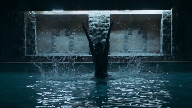 Night footage of beautiful young girl with amazing athletic sexy body in black bikini jumps two times in the swimming pool with water splashes, slow motion movie shooting of female model like a ghost
