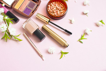 Various cosmetic products eye shadow powder lip gloss blush mascara brush for makeup and white spring flowers in a white gift bag on a pink pastel background top view with copy space