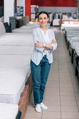 smiling female customer with arms crossed standing in furniture store with arranged mattresses