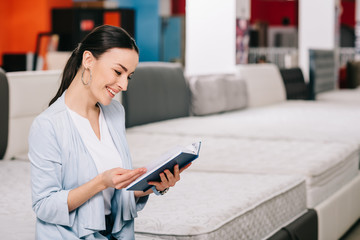 side view of smiling female customer with notebook in furniture store with arranged mattresses