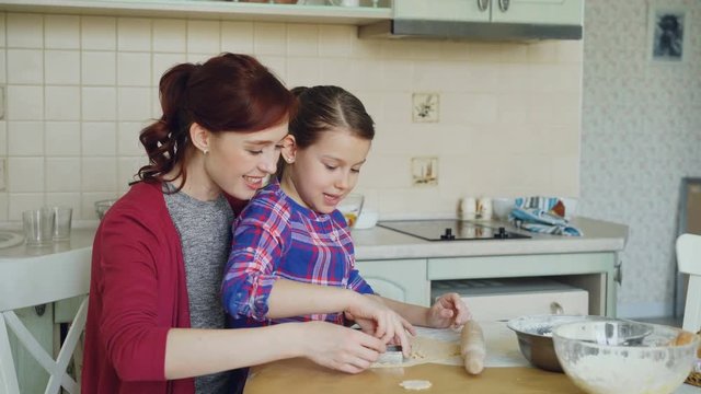 Smiling mother and cute daughter making cookies together using bakery forms while sitting in modern kitchen at home. Family, food and people concept