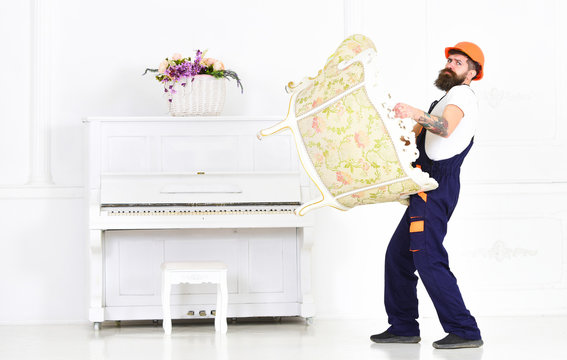Man With Beard, Worker In Overalls And Helmet Lifts Up Armchair, White Background. Courier Delivers Furniture In Case Of Move Out, Relocation. Loader Carries Armchair. Delivery Service Concept.