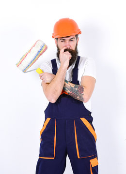Renovation and decoration concept. Man with beard and mustache in helmet and overalls with paint roller. Decorator on serious face holds paint roller and puts finger into nostril, white background.