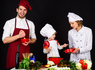 Happy family in kitchen. Healthy food at home. Adorable kid in chef hat. Preparation to dinner. Parents with child preparing dinner. Cheerful parents teaching a boy how to cook. Homemade food.