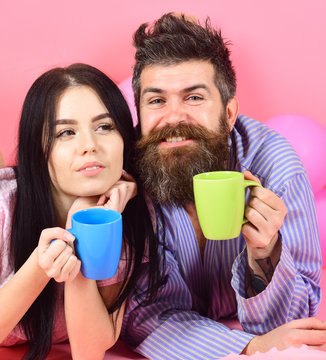 Man and woman on smiling faces lay, pink background. Couple in love drink coffee in bed. Couple relax in morning with coffee. Weekend morning concept. Man and woman in domestic clothes, pajamas.
