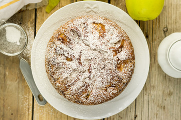 HOMEMADE RUSTIC APPLE CAKE PIE POWDERED WITH SUGAR ON A WHITE PLATE ON A WOODEN TABLE WITH FRESH APPLE ON A SIDE