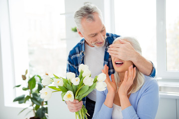 Portrait of lovely sweet attractive cheerful couple in casual outfit celebrating anniversary, man closing her wondered, amazed lover eyes with palm preparing bouquet of tulips