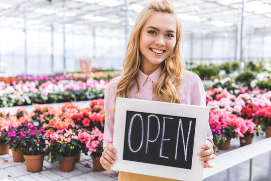 Attractive female gardener holding Open board by flowers in glasshouse - Powered by Adobe