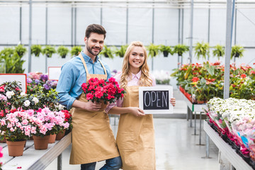 Fototapeta premium Young man and blonde woman holding Open board by flowers in glasshouse