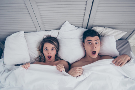 What we did last night? Top view of funny man and woman lying in bed covered by sheets with wide opened eyes and mouth looking at camera, having shock, confuse