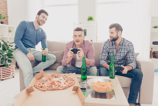 Three attractive handsome stylish men in casual outfits playing videogame, laughing from frustrated upset loser sitting in living room, drinking  beer, lager from bottle, having pizza, chips