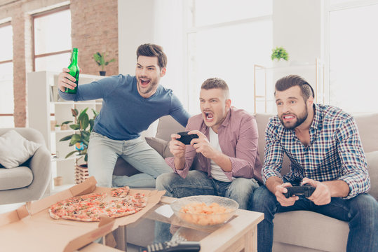 Portrait of stylish attractive modern successful three men in casual outfits playing video game holding joystick in hands, having snack, pizza, chips, good, alcohol beverages on the table