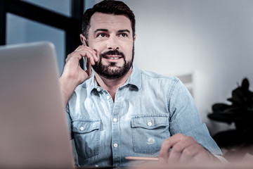 Best connection. Satisfied handsome busy man sitting by the table near the laptop looking aside and holding cellphone near his ear.