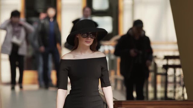Stylish attractive lady in black dress, hat and sunglasses walking down the street in the Mall. Classic fashion concept. Slow motion