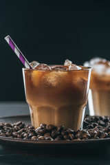 selective focus of glass of cold iced coffee with straw on plate with roasted coffee beans on tabletop on dark backdrop