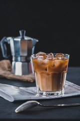 selective focus of glass of cold iced coffee, newspaper and coffee maker on tabletop on dark backdrop