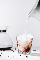 close up view of pouring milk into glass of cold brewed coffee process, coffee maker and roasted coffee beans on grey background