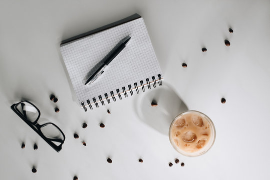 Flat Lay With Glass Of Cold Iced Coffee, Roasted Coffee Beans, Eyeglasses, Empty Notebook And Pen On White Surface