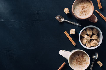 top view of cups of coffee, spoons, bowl of cane sugar and cinnamon stick on dark tabletop