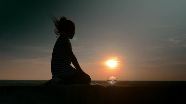 beautiful idyllic scene sitting little kid girl's silhouette and round aquarium floating fish outdoors on backdrop of the sea at sunset in summer