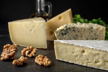 Different kinds of cheeses on black stone board with nuts and herbs.