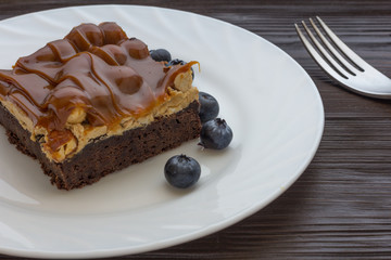 Brownie cake with caramel, nougat, nuts and fresh blueberries in white plate with fork on brown wood background