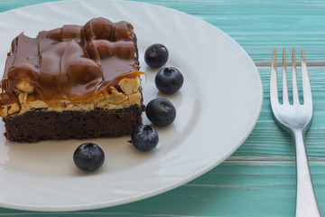 Brownie cake with caramel, nougat, nuts and fresh blueberries in white plate with fork on blue backgound