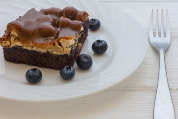 Brownie cake with caramel, nougat, nuts and fresh blueberries in white plate with fork on white background