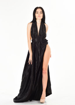 Woman in elegant black long evening dress with decollete, white background. Attractive girl wears expensive fashionable evening dress with erotic slit. Lady, sexy girl in dress. Fashion dress concept.