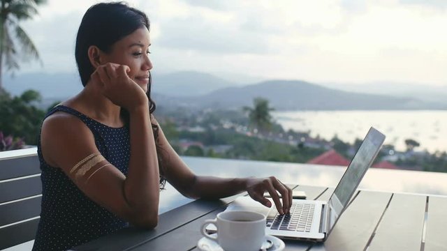 Beautiful asian woman using laptop for typing message in social media. Happy business woman working on notebook computer on summer terrace with panoramic sea landscape on luxury villa