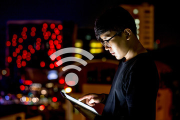 Glasses Asian man look at his computer tablet during the night with city lights. Focus at his hand. Shallow depth of field. There are wifi icon Floating on air.