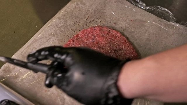 Cook makes meat medallions for burgers. Chef in black food gloves makes cutlet. Cutlets are leveled in steel ring in an even medallion. On top of pouring spices pepper and salt. Meat marbled beef lies