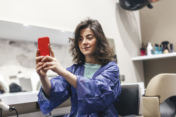 Young woman takes selfie with her smartphone in front of the mirror of a hairdresser's shop after cutting and styling