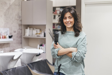 Professional hairdresser with hairbrushes and scissors in hand, the young woman standing in salon with folded hands and smiling