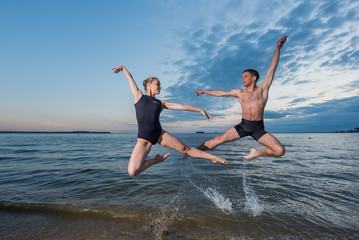 Young guy and girl in black swimwear high jump and dance on the beach in the evening. Street ballet.