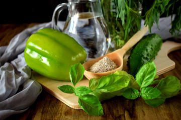 A green pepper, brans in a wooden spoon, basil, tarragon, a cucumber and water in a glass jug. Ingredients for preparing a healthy meal