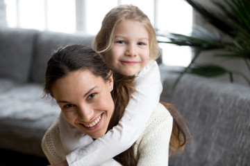 Happy single mother laughing piggybacking little girl at home, smiling mom and daughter having fun...