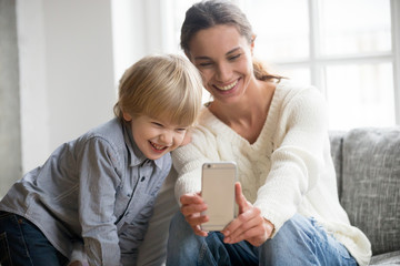 Happy mother laughing taking selfie with little son on smartphone at home, smiling single mom and cute adopted boy playing making photo posing for self portrait, mommy and kid watching video on cell