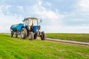 Obraz premium Agriculture, a tractor with a barrel trailer rides along the green field.