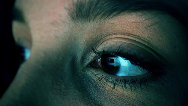 Extreme closeup of caucasian woman surfing internet at night, using a device before sleep with insomnia or sleeplessness. Screen with social network reflecting in her eyes. Internet addiction concept